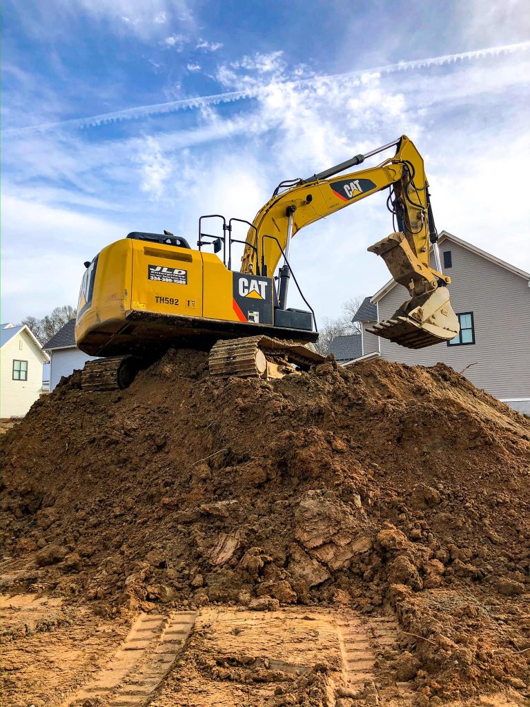 Yellow excavator on a dirt mound in a construction site, Auburn, AL, with houses and blue sky in the background. Grading.